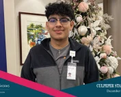 A young man (Joel) with glasses and curly hair smiles while standing in front of a decorated Christmas tree. Hes wearing a badge and dark sweatshirt. The image features Virginia Career Works and Culpeper Star-Exponent branding.