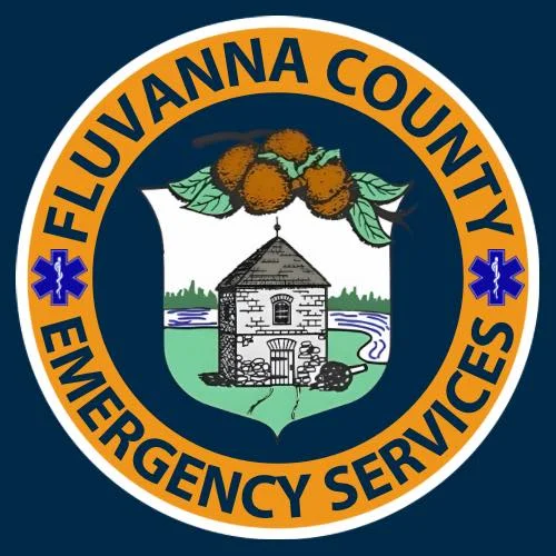 Fluvanna EMS Circular logo for Fluvanna County Emergency Services, featuring a central shield with a stone building, trees, and a river, surrounded by an orange ring with blue text and two blue medical symbols.