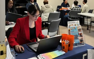 A woman in a red blazer works on a laptop at a table with brochures and orange cups, while several people sit and talk in groups in a classroom-like setting. The tablecloth reads “Pathways into Careers.”.