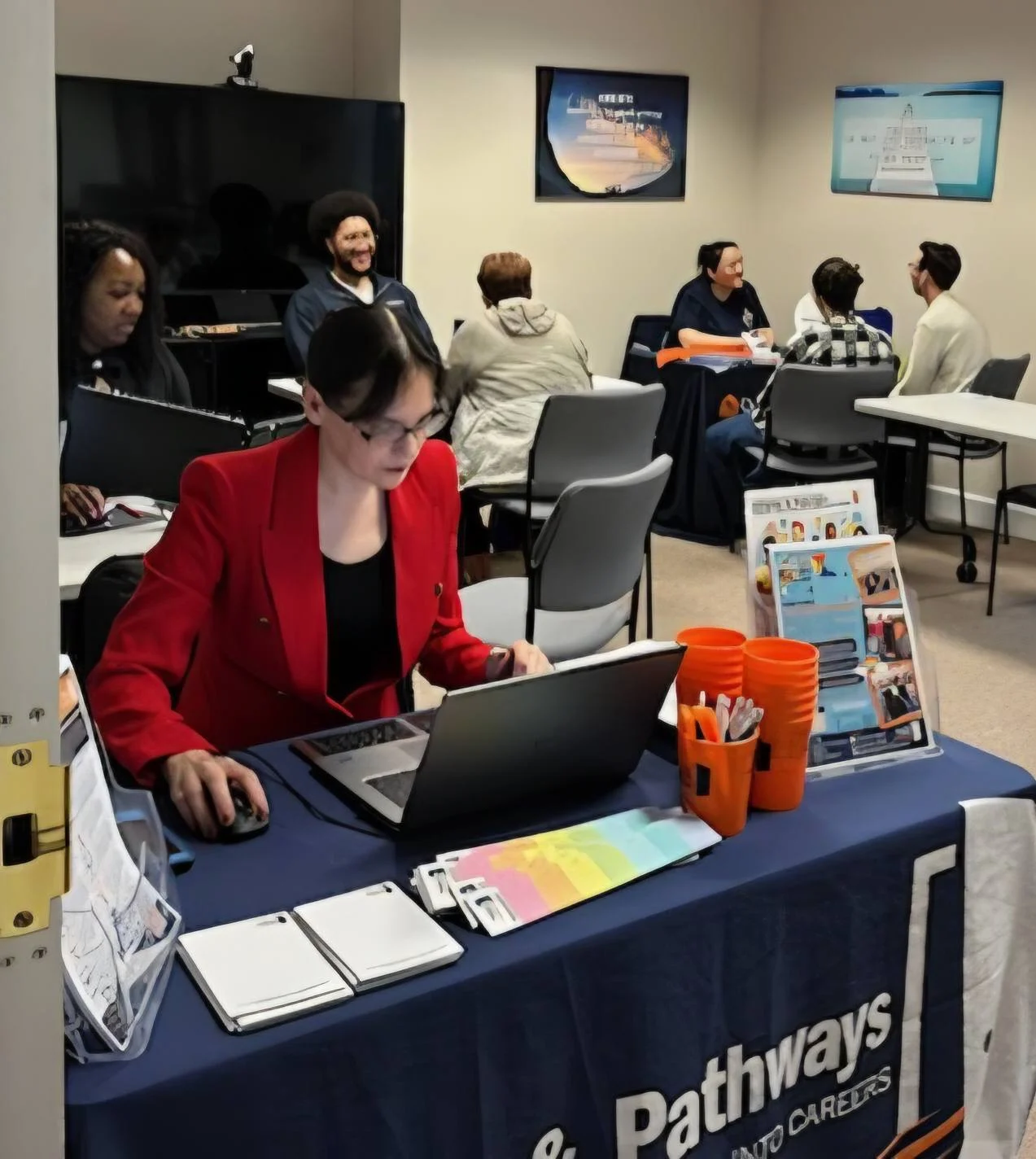 A woman in a red blazer works on a laptop at a table with brochures and orange cups, while several people sit and talk in groups in a classroom-like setting. The tablecloth reads “Pathways into Careers.”.